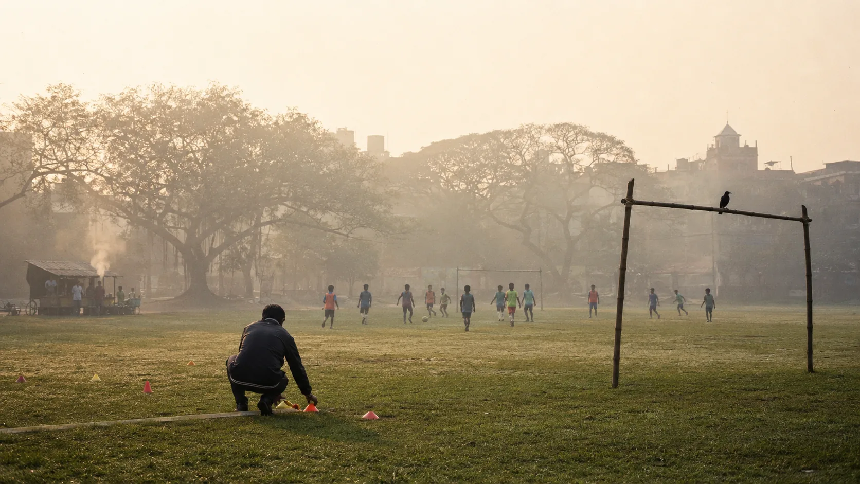 A coach lays out training cones on a misted Kolkata ground at dawn while children play in the distance.